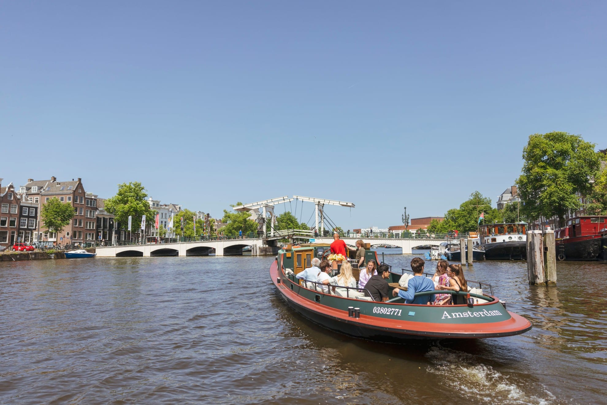 Un bateau de croisière classique rempli de passagers navigue sur un canal d'Amsterdam en direction du Magere Brug par une journée ensoleillée.